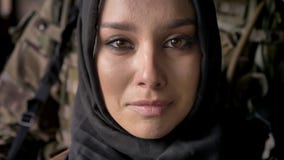 Nära stående av den unga muslimkvinnan, i gråt och att se för hijab kameran, beväpnad soldat med vapenanseende bakom lager videofilmer Nära stående av den unga muslimkvinnan, i gråt och att se för hijab kameran, beväpnad soldat med vapenanseende bakom lager videofilmer