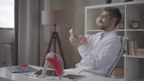 O doutor farpado bem sucedido, satisfeito feliz dispersa os dólares que sentam-se em uma cadeira em uma tabela de trabalho na clí video estoque O doutor farpado bem sucedido, satisfeito feliz dispersa os dólares que sentam-se em uma cadeira em uma tabela de trabalho na clí video estoque