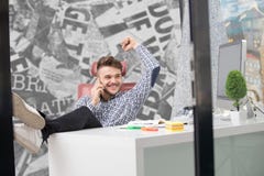 O homem de negócios considerável na camisa clássica está usando um smartphone e está sorrindo ao sentar-se com pés na tabela no e foto de stock O homem de negócios considerável na camisa clássica está usando um smartphone e está sorrindo ao sentar-se com pés na tabela no e foto de stock