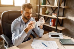 O homem de negócios doente novo senta-se na tabela em seu próprio escritório Guarda o guardanapo e o copo brancos nas mãos O indi fotos de stock royalty free O homem de negócios doente novo senta-se na tabela em seu próprio escritório Guarda o guardanapo e o copo brancos nas mãos O indi fotos de stock royalty free
