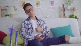 O homem novo asiático alegre com um pé quebrado nas ataduras usa um smartphone que olha a câmera e que mostra os polegares acima video estoque O homem novo asiático alegre com um pé quebrado nas ataduras usa um smartphone que olha a câmera e que mostra os polegares acima video estoque