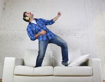 o homem 20s ou 30s saltou no sofá que escuta a música no telefone celular com os fones de ouvido que jogam Air Guitar imagem de stock royalty free o homem 20s ou 30s saltou no sofá que escuta a música no telefone celular com os fones de ouvido que jogam Air Guitar imagem de stock royalty free