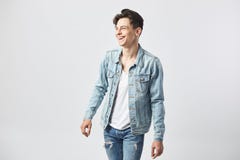 O indivíduo de cabelo escuro novo de sorriso em um t-shirt branco, em calças de brim e em um revestimento da sarja de Nimes está fotografia de stock O indivíduo de cabelo escuro novo de sorriso em um t-shirt branco, em calças de brim e em um revestimento da sarja de Nimes está fotografia de stock