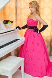 O louro elegante no vestido vermelho longo está estando ao lado do piano fotografia de stock royalty free O louro elegante no vestido vermelho longo está estando ao lado do piano fotografia de stock royalty free