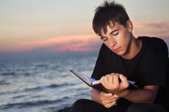 O menino do adolescente lê o livro que senta-se na praia imagem de stock O menino do adolescente lê o livro que senta-se na praia imagem de stock