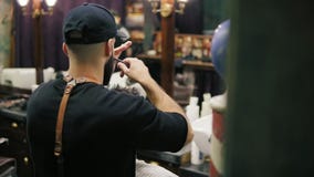 Opinião lateral e traseira o barbeiro à moda com a barba que executa um corte de cabelo com as tesouras que guardam uma escova de video estoque Opinião lateral e traseira o barbeiro à moda com a barba que executa um corte de cabelo com as tesouras que guardam uma escova de video estoque