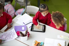 Opinión elevada dos colegialas infantiles que se sientan en una tabla, usando una tableta y una aguja en una sala de clase fotos de archivo Opinión elevada dos colegialas infantiles que se sientan en una tabla, usando una tableta y una aguja en una sala de clase fotos de archivo