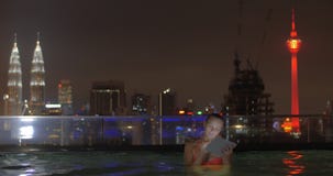 Opinión la mujer en piscina en el tejado del rascacielos usando la tableta contra paisaje de la ciudad de la noche Kuala Lumpur, almacen de metraje de vídeo Opinión la mujer en piscina en el tejado del rascacielos usando la tableta contra paisaje de la ciudad de la noche Kuala Lumpur, almacen de metraje de vídeo
