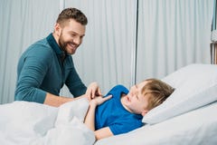 Pai de sorriso que joga com o rapaz pequeno doente que encontra-se na cama de hospital foto de stock Pai de sorriso que joga com o rapaz pequeno doente que encontra-se na cama de hospital foto de stock