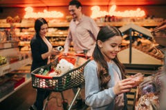 Pais e filha novos na mercearia Caixa pequena alegre da posse da menina com filhós e sorriso Os pais estão atrás em fotografia de stock Pais e filha novos na mercearia Caixa pequena alegre da posse da menina com filhós e sorriso Os pais estão atrás em fotografia de stock