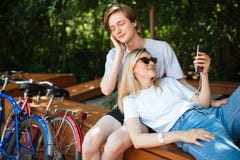 Pares frescos novos na música de escuta dos fones de ouvido no parque com duas bicicletas próximo Menino que senta-se no banco e foto de stock Pares frescos novos na música de escuta dos fones de ouvido no parque com duas bicicletas próximo Menino que senta-se no banco e foto de stock