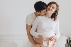 Pares grávidos novos felizes guardando brancos poucas peúgas na colisão da barriga na cama branca Família, mamã e paizinho grávid imagens de stock Pares grávidos novos felizes guardando brancos poucas peúgas na colisão da barriga na cama branca Família, mamã e paizinho grávid imagens de stock