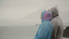 Pares novos nas capas de chuva que estão na lagoa do gelo em Islândia e que mostram algo a cada outro com dedos video estoque Pares novos nas capas de chuva que estão na lagoa do gelo em Islândia e que mostram algo a cada outro com dedos video estoque
