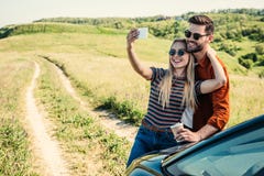pares à moda de sorriso nos óculos de sol com o copo de café que toma o selfie no smartphone perto do carro em rural imagens de stock pares à moda de sorriso nos óculos de sol com o copo de café que toma o selfie no smartphone perto do carro em rural imagens de stock