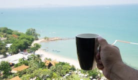 Passi la tazza di caffè della tenuta con la spiaggia nel fondo immagini stock libere da diritti Passi la tazza di caffè della tenuta con la spiaggia nel fondo immagini stock libere da diritti