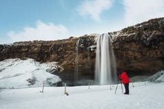 Paysage de cascade d'hiver, photographe dans la veste rouge prenant la photographie avec le trépied d'appareil-photo à la cascade photo libre de droits Paysage de cascade d'hiver, photographe dans la veste rouge prenant la photographie avec le trépied d'appareil-photo à la cascade photo libre de droits
