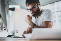 Pensive bearded man wearing eye glasses and white tshirt, working at modern loft studio-office.Blurred background stock photos Pensive bearded man wearing eye glasses and white tshirt, working at modern loft studio-office.Blurred background stock photos