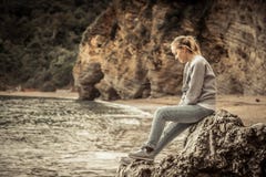 Pensive lonely young woman traveler relaxing on a big cliff stone on the beach looking at wild mountain scenery in retro vintage s stock photography Pensive lonely young woman traveler relaxing on a big cliff stone on the beach looking at wild mountain scenery in retro vintage s stock photography