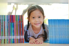 Pequeña muchacha asiática feliz del niño que miente en el estante en la biblioteca Creatividad de los niños y concepto de la imag fotografía de archivo libre de regalías Pequeña muchacha asiática feliz del niño que miente en el estante en la biblioteca Creatividad de los niños y concepto de la imag fotografía de archivo libre de regalías