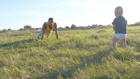 Pequeño bebé que se coloca en hierba verde en el prado y que mira como su papá que hace pectorales Hombre atlético que hace pecto fotos de archivo Pequeño bebé que se coloca en hierba verde en el prado y que mira como su papá que hace pectorales Hombre atlético que hace pecto fotos de archivo