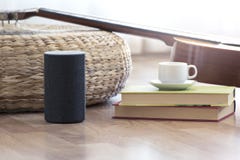 Personal assistant loudspeaker on a wooden floor of a smart home living room. Next, a guitar and some books and a cup of coffee.
