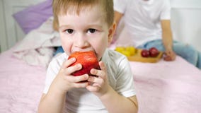 Peu de gar?on mange la pomme dans la chambre ? coucher du parent Ses parents s'asseyant sur le lit sur le fond images stock Peu de gar?on mange la pomme dans la chambre ? coucher du parent Ses parents s'asseyant sur le lit sur le fond images stock