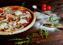 A pizza caseiro do presunto, do salame e do cogumelo serviu em uma placa em uma mesa de cozinha de madeira rústica velha cercada foto de stock A pizza caseiro do presunto, do salame e do cogumelo serviu em uma placa em uma mesa de cozinha de madeira rústica velha cercada foto de stock