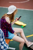 Piękna blond dziewczyna jest ubranym w kratkę koszula, nakrętkę i drelichowych skróty, siedzi na sporta polu z telefonem w ona obrazy royalty free Piękna blond dziewczyna jest ubranym w kratkę koszula, nakrętkę i drelichowych skróty, siedzi na sporta polu z telefonem w ona obrazy royalty free