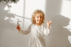 Playful cheerful little beautiful girl with blonde curly hair plays with toy deer, dressed in festive white dress, ready stock photo Playful cheerful little beautiful girl with blonde curly hair plays with toy deer, dressed in festive white dress, ready stock photo