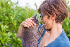 Playful domestic cat held and cuddled by smiling woman with eyeglasses. Outdoor setting in green home garden. Shallow depth of fie stock image Playful domestic cat held and cuddled by smiling woman with eyeglasses. Outdoor setting in green home garden. Shallow depth of fie stock image