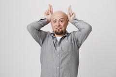 Playful positive bald male with beard has cheerful expression, wears gray shirt, behaves as rabbit, holds fingers above Royalty Free Stock Images Playful positive bald male with beard has cheerful expression, wears gray shirt, behaves as rabbit, holds fingers above Royalty Free Stock Images