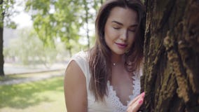 Portrait adorable young girl with long brunette hair wearing a long white summer fashion dress standing next to a tree stock footage Portrait adorable young girl with long brunette hair wearing a long white summer fashion dress standing next to a tree stock footage