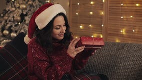 Portrait of attractive young brunette woman with wavy hair in santas hat sitting near Christmas tree at grey sofa stock video Portrait of attractive young brunette woman with wavy hair in santas hat sitting near Christmas tree at grey sofa stock video