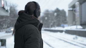 Portrait d'un jeune homme aux cheveux longs avec une barbe dans des écouteurs se tenant à un arrêt de tram en hiver et attendant banque de vidéos Portrait d'un jeune homme aux cheveux longs avec une barbe dans des écouteurs se tenant à un arrêt de tram en hiver et attendant banque de vidéos