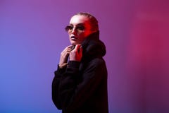 Portrait de fille de mode dans un chandail noir avec un capot et des lunettes de soleil autour de forme dans la lampe au n?on dan images stock Portrait de fille de mode dans un chandail noir avec un capot et des lunettes de soleil autour de forme dans la lampe au n?on dan images stock