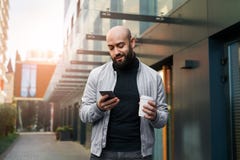 Portrait de jeune homme de sourire utilisant le smartphone sur la rue de ville L'homme envoie le message textuel, boit du café li image stock Portrait de jeune homme de sourire utilisant le smartphone sur la rue de ville L'homme envoie le message textuel, boit du café li image stock