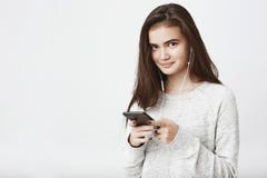 Portrait de modèle européen beau gai avec de longs cheveux bruns, tenant le smartphone tout en souriant largement et images libres de droits Portrait de modèle européen beau gai avec de longs cheveux bruns, tenant le smartphone tout en souriant largement et images libres de droits
