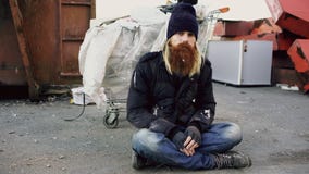 Portrait du jeune homme sans abri barbu s'asseyant sur un trottoir près du récipient de déchets d'ANG de caddie pendant l'hiver f photographie stock Portrait du jeune homme sans abri barbu s'asseyant sur un trottoir près du récipient de déchets d'ANG de caddie pendant l'hiver f photographie stock