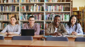 Portrait of four multi-ethnic students sitting at long desk in big spacious library with piles of books looking at stock video Portrait of four multi-ethnic students sitting at long desk in big spacious library with piles of books looking at stock video