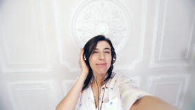 Portrait of funny young woman, girl, brunette, in white shirt, in headphones, she listening to music, shoots video stock video footage Portrait of funny young woman, girl, brunette, in white shirt, in headphones, she listening to music, shoots video stock video footage