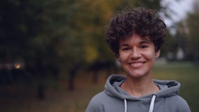 Portrait of good-looking girl with short curly hair looking at camera smiling then laughing standing outdoors in autumn stock footage Portrait of good-looking girl with short curly hair looking at camera smiling then laughing standing outdoors in autumn stock footage