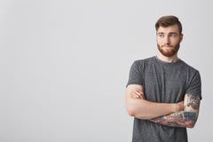 Portrait of handsome bearded hipster guy with dark hair and tattoo sleeve looking aside with crossed hands and cynical Stock Photo Portrait of handsome bearded hipster guy with dark hair and tattoo sleeve looking aside with crossed hands and cynical Stock Photo