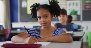 Portrait of happy mixed race schoolgirl sitting at classroom, making notes, looking at camera