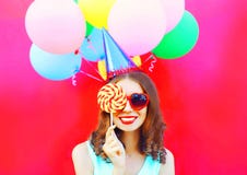 Portrait happy smiling woman in a birthday cap closes her eye with a lollipop on stick over an air colorful balloons on pink bac stock photo Portrait happy smiling woman in a birthday cap closes her eye with a lollipop on stick over an air colorful balloons on pink bac stock photo