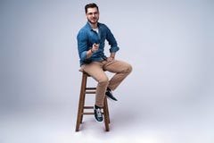 Portrait intégral d'un jeune homme attirant dans la chemise de jeans se reposant sur la chaise au-dessus du fond gris photographie stock Portrait intégral d'un jeune homme attirant dans la chemise de jeans se reposant sur la chaise au-dessus du fond gris photographie stock