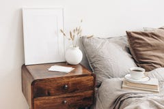 Portrait white frame mockup on retro wooden bedside table. Modern white ceramic vase, dry Lagurus ovatus grass. Cup of