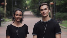 Portrait of Young Caucasian Man and Muslin Woman Wearing Headphones Looking Happy Standing in the Park Turning Heads to stock video footage Portrait of Young Caucasian Man and Muslin Woman Wearing Headphones Looking Happy Standing in the Park Turning Heads to stock video footage