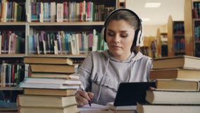 Positive junge lächelnde Frau in den weißen Kopfhörern sitzt bei Tisch in der digitalen Tablette des Universitätsbibliothekbiblio stock video Positive junge lächelnde Frau in den weißen Kopfhörern sitzt bei Tisch in der digitalen Tablette des Universitätsbibliothekbiblio stock video