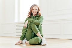 Positive redhead fitness girl feels relaxed, wears tracksuit, sportshoes, has rest after yoga training indoor, poses on floor, royalty free stock photo Positive redhead fitness girl feels relaxed, wears tracksuit, sportshoes, has rest after yoga training indoor, poses on floor, royalty free stock photo