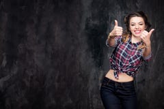 Pouces de signe d'exposition de fille bons dans la chemise à carreaux et des jeans sur le fond gris-foncé photos libres de droits Pouces de signe d'exposition de fille bons dans la chemise à carreaux et des jeans sur le fond gris-foncé photos libres de droits